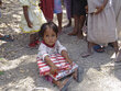 A young child sits on a dirt ground holding a pack of fortified biscuits, surrounded by several other children standing barefoot nearby outdoors.