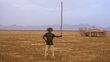 A man holding a big stick up high stands in the middle of dry land with a hut and hills behind. Photo: UNEP/Malachie Nabbi