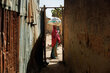 A girl carries a WFP food sack on her head while walking through a narrow passageway between buildings.