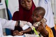 A closeup of a health worker's hand holding a MUAC nutrition band around a little girl's arm that shows her testing positive for moderate-acute malnutrition, as her mother holds the child. Photo: WFP/Sylvie Njobati
