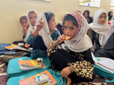 Afghan school girls wearing long robes and headscarves sit on a rug with backpacks and school books in front of them,  eating WFP-provided high-energy biscuits in Afghanistan.  Photo: WFP/Isheeta Sumra