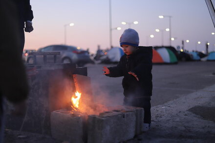 Child in a hat warms hands over a small fire made from bricks at a roadside camp, with tents and cars visible in the background at dusk.