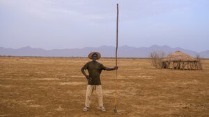 A man holding a big stick up high stands in the middle of dry land with a hut and hills behind. Photo: UNEP/Malachie Nabbi