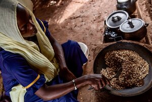 A woman in a blue dress and yellow headscarf sits on an earth floor inside stirring a bowl of grains. Photo: WFP/Gabriela Vivacqua