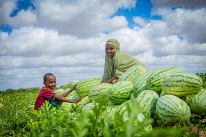 A smiling boy and woman sit in a green field surrounded by watermelons. Photo: WFP/William Orlale