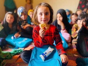 A smiling little girl in a red top sits in an Afghan classrooms holding up a WFP fortified biscuit. Photo: WFP/Sayed Abidullah Sadat