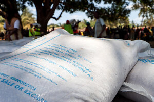 Copilot said:  WFP food assistance sacks stacked at a distribution site as people gather beneath trees in the background, awaiting humanitarian support.