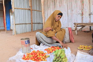 A woman arranges tomatoes and green bananas for sale outside a corrugated metal shelter, with produce displayed on a cloth in a sandy area.