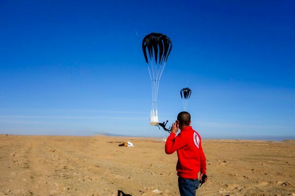 man in red jacket watching as black parachutes land on sandy ground