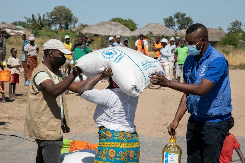 Two men in WFP uniforms assist a woman with carrying a bag which has a WFP logo on it.
