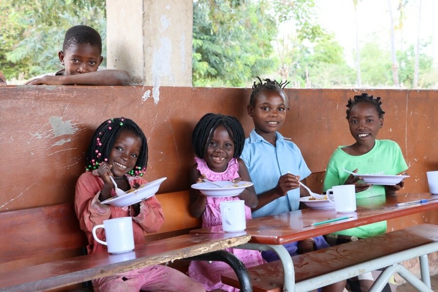 Four children smile and enjoy food while one child smiles standing behind them.