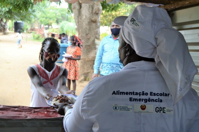 A girl wearing a red patterned mask receives a plate of food from a woman in white.