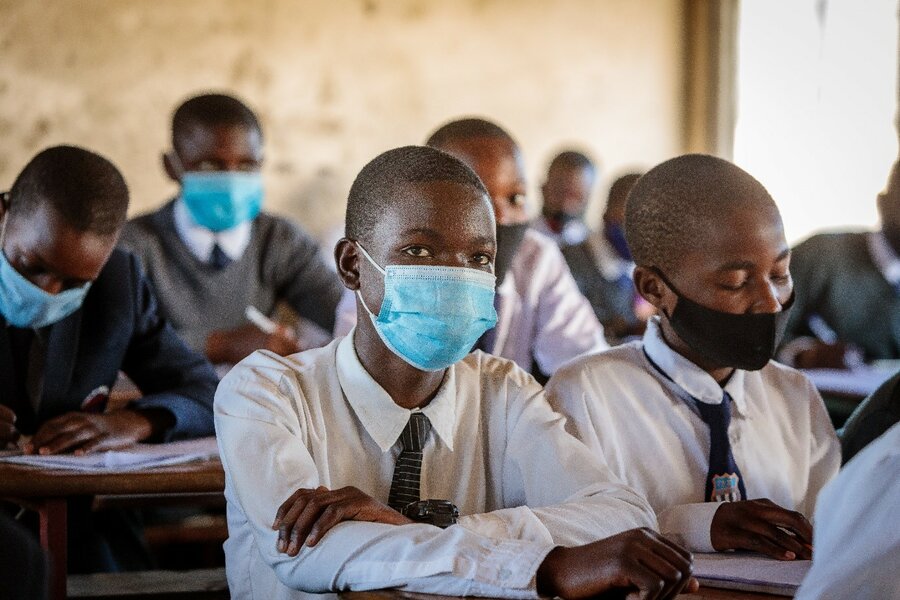 A boy wearing a blue mask sits in a classroom surrounded by peers.
