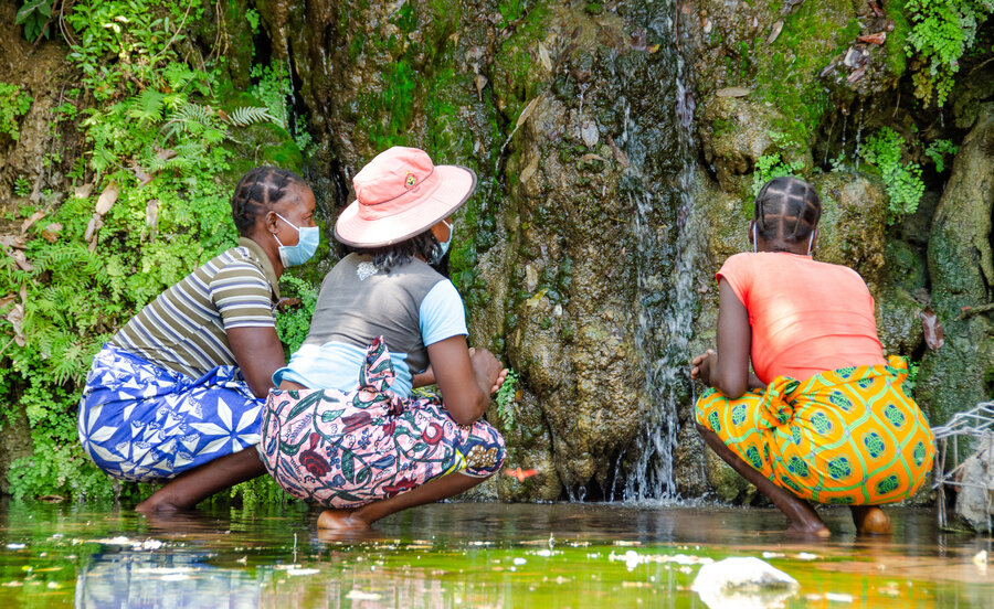 Three women crouch in shallow water looking at a rock.
