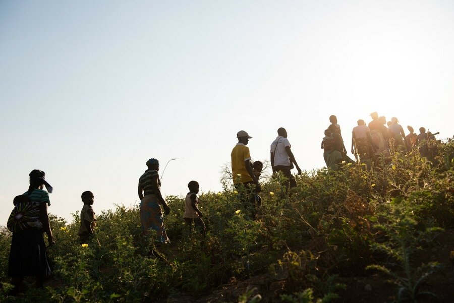 A group of men, women and children walk in a line up a hill.
