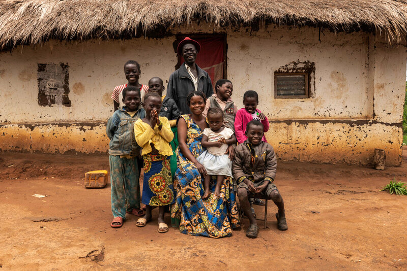 A group of ten people pose in front of a thatched roof home.