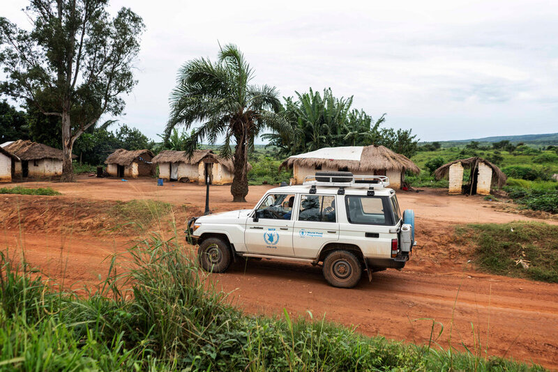 A WFP vehicle driving on brown land in front of buildings with thatched roofs.