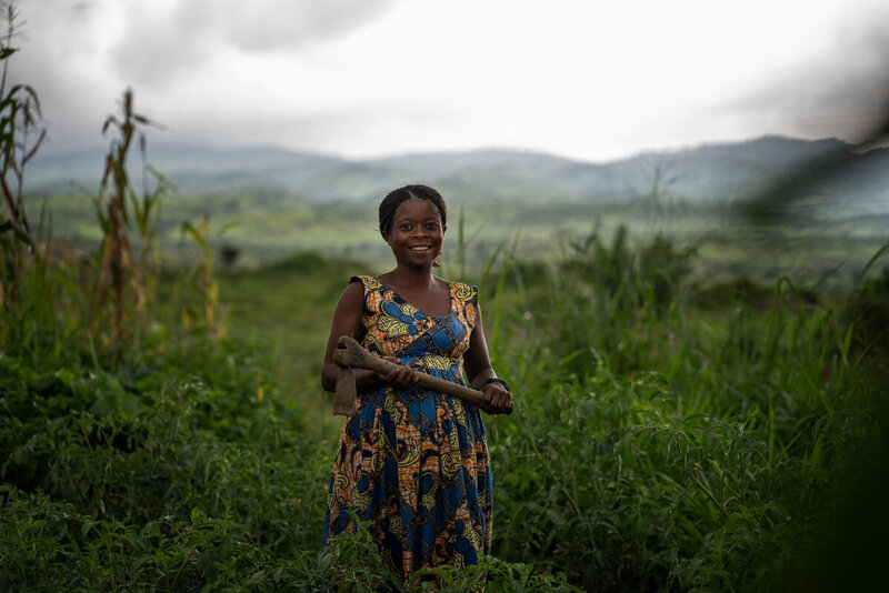 A woman in a blue patterned dress smiles to the camera with a green field and hills behind her.