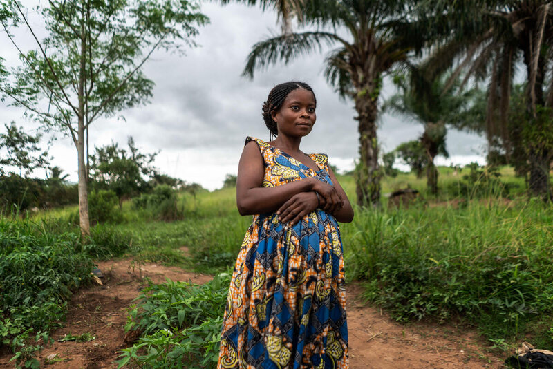 A woman in a blue patterned dress looks to the camera with a green field behind her.