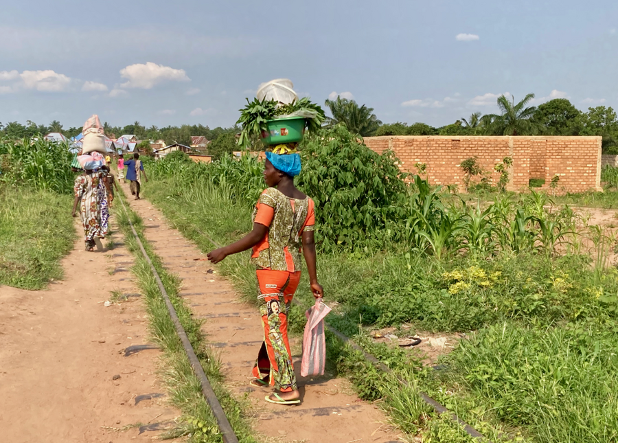 Women carry products on their head while walking on dirt roads.