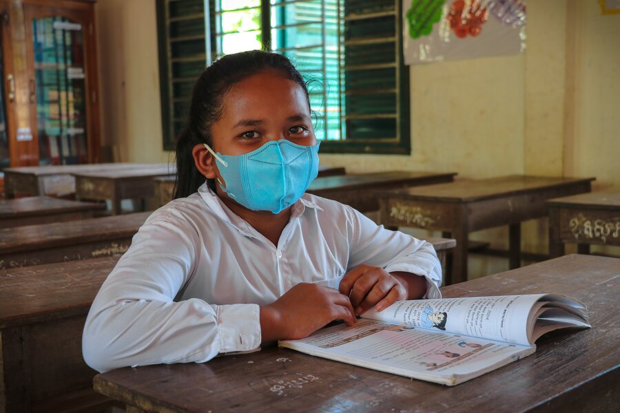 A girl wearing a mask sits in a classroom in Cambodia with a book open on the table in front of her. 