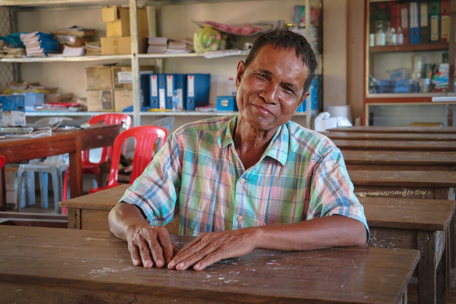 A man sits in a classroom while smiling at the camera and tilting his head