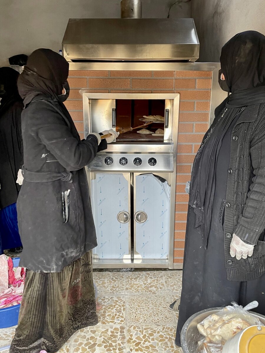 Two women place bread inside an oven.