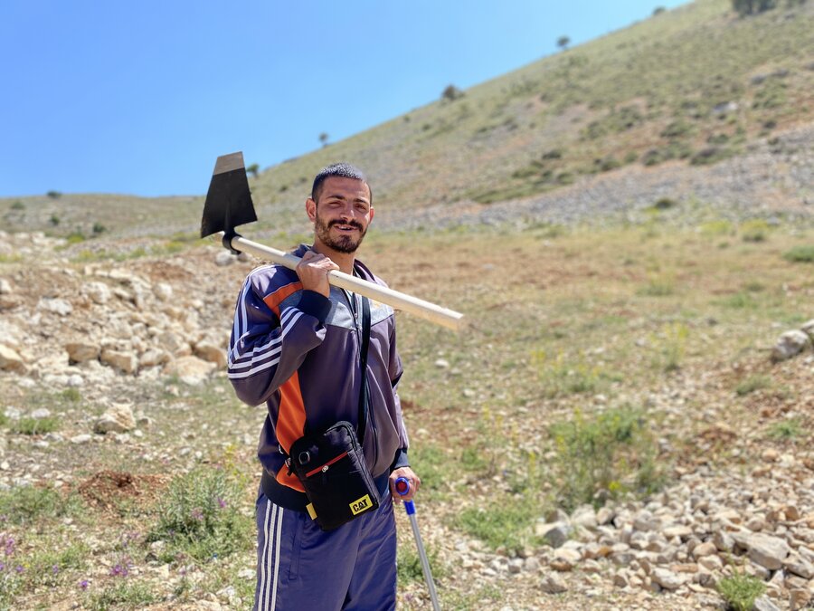 A farmer holds a farming tool over his shoulder and looks at the camera