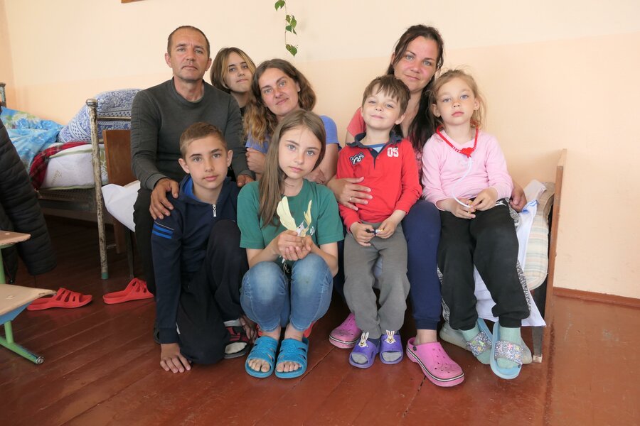 A family of 2 adults and 6 children sit closely together on a bed in their temporary accommodation