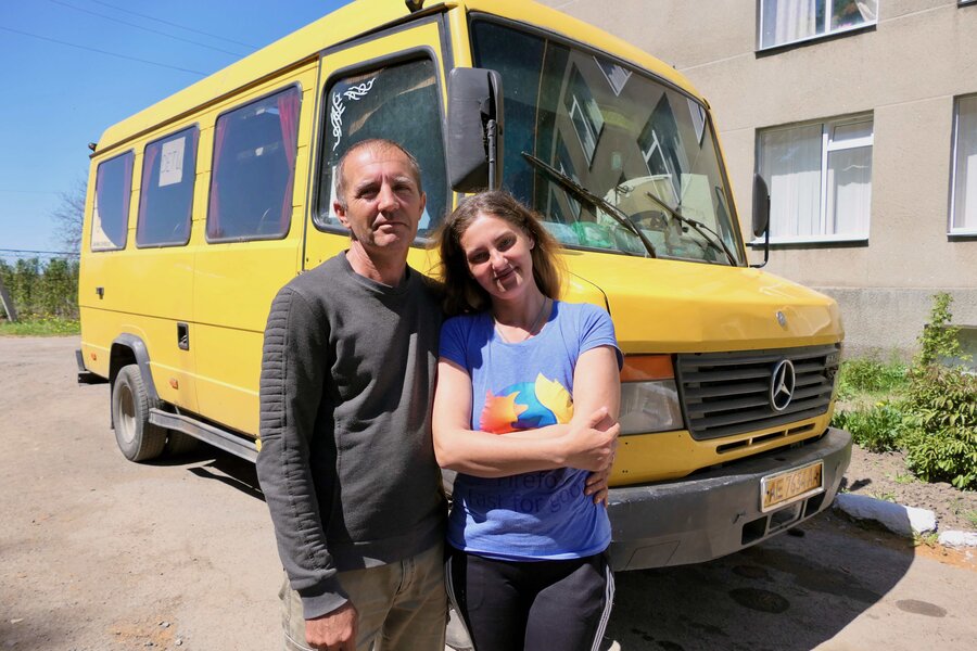 2 adults stand together in front of a yellow bus