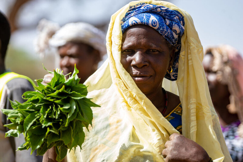 A women wearing a yellow headscarf holds some green vegetables while looking at the camera