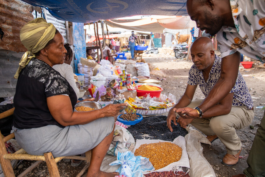 A woman sits and sells food in a market to 2 male shoppers