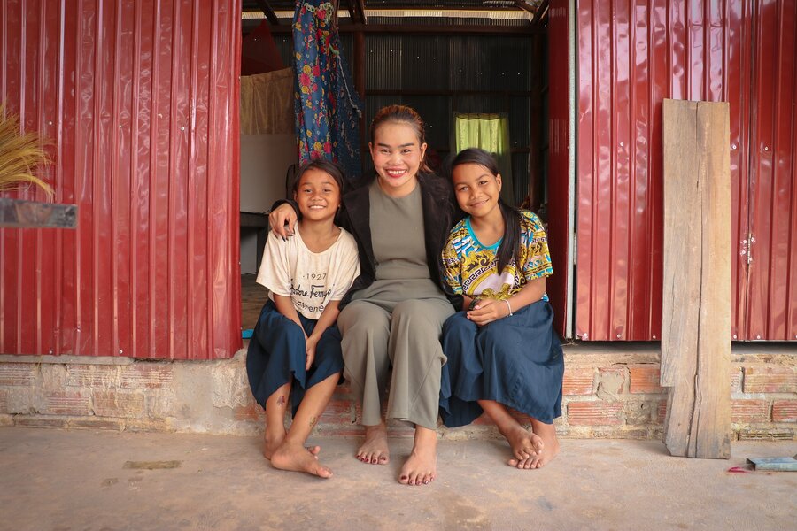 A mother and her 2 daughters sit with their arms around each other in the doorway to their home