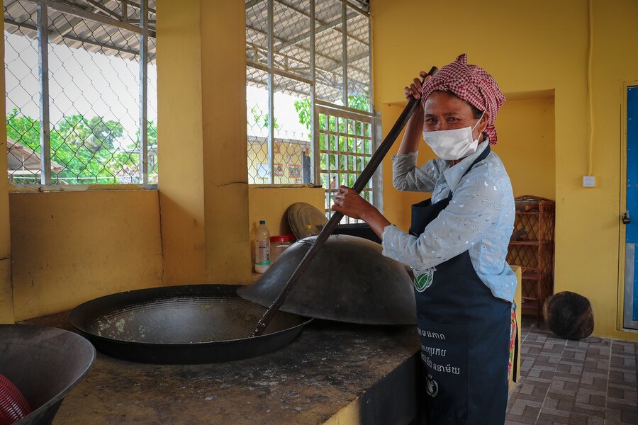 A female cook in a school holds a long spoon to stir a large pot of food.