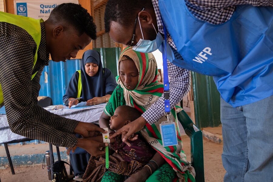 A mother holds a baby while they are checked for malnutrition by 2 medical staff 