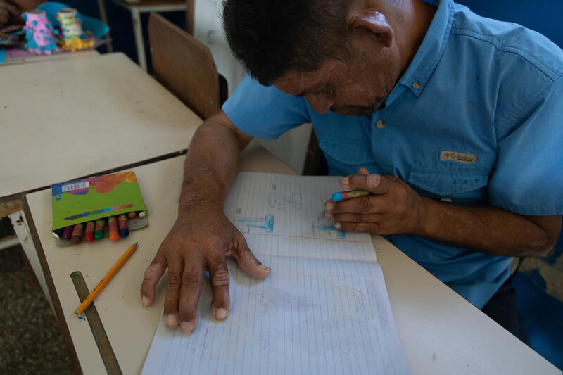 A man in blue shirt sits at a school desk writing out numbers