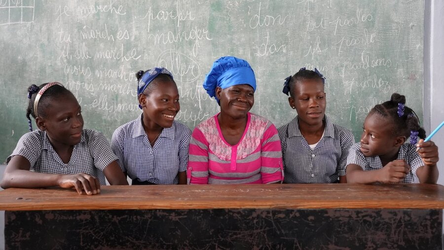 Haitian woman in pink shirt and blue headdress sits with four girls in school uniforms against a blackboard