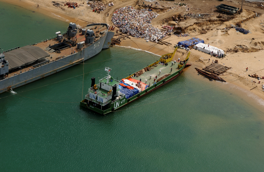 Aerial shot of two vessels, one a UN/WFP landing craft on a beach where aid is assembled