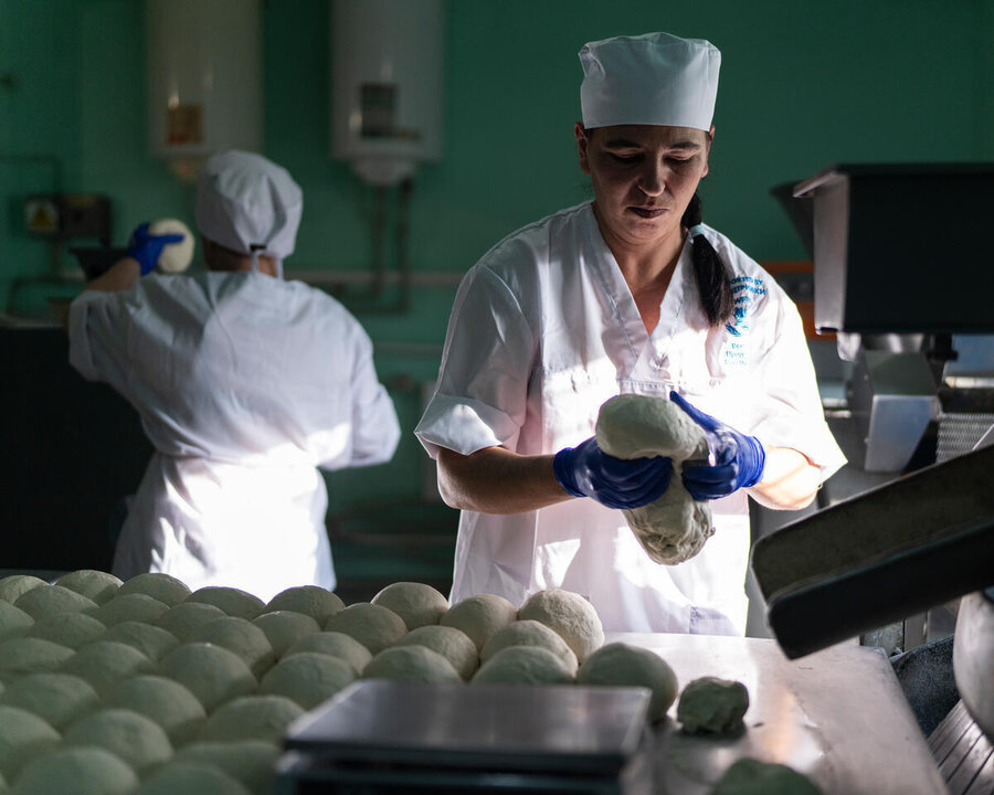 A baker in white uniform kneads dough, another baker back to the camer in the background