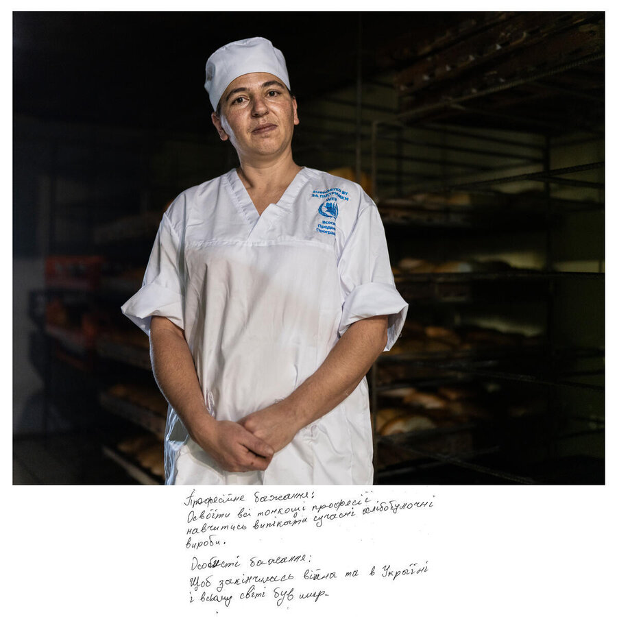 A woman in white overalls, with a WFP logo and bakers hat in front of a dark background where rows of freshly baked bread can be discerned