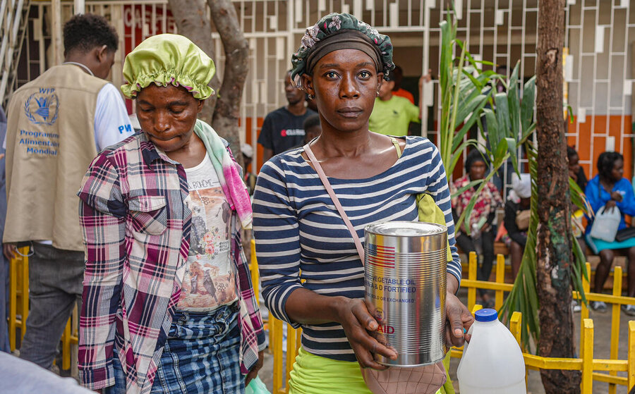 A man with his back to the camera, WFP jacket, two women in foreground, one carrying a can of food assistance