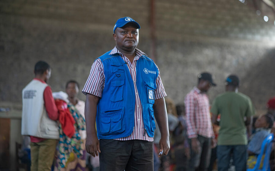 A Black man wearing a blue cap and a blue WFP vest looks into the camera, people blurred in background