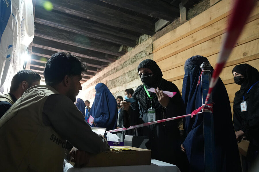 A man sits at a cash distribution desk as his female colleagues checks the ID of a woman