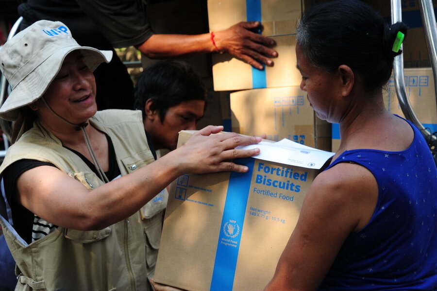 A woman in a soft white hat hands a box of WFP-branded fortified biscuits to a woman in a blue vest top. A man and other boxes are visible in the background.