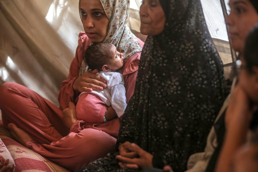 Person in pink outfit sits cross-legged holding a baby in white and pink; another person in dark patterned clothing is partially visible on the right.