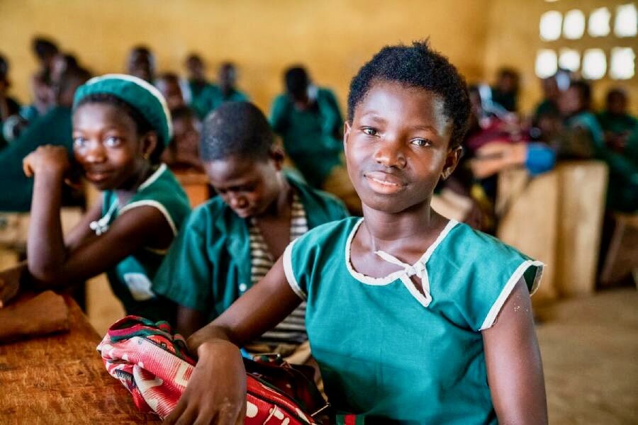 A smiling young schoolgirl wearing a green uniform sits in a classroom with other students. Photo: WFP/Michael Duff