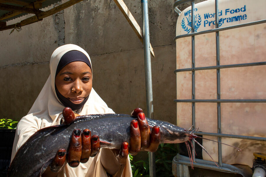 A young woman in Islamic dress holds up a big fish with a WFP sign in the background