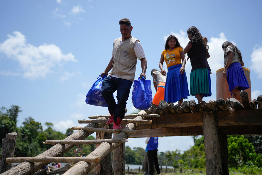 A man in a WFP jacket carries school meals in blue bags as he steps down from a wooden jetty