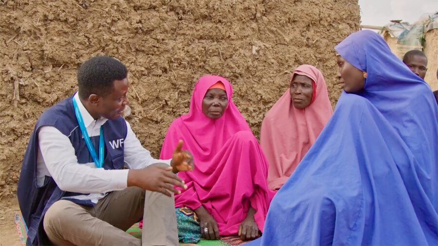 A WFP staffer, wearing a blue WFP vest and white shirt, sits near a mud-brick building, chatting with displaced women wearing colourful gowns in northeastern Nigeria. Photo: WFP/Arete/Damilola Onafuwa