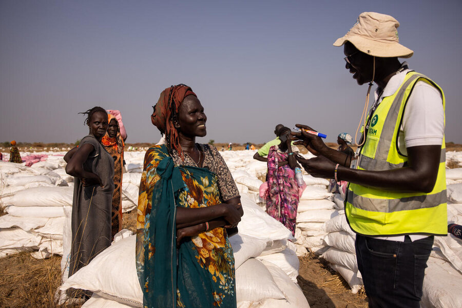 A WFP staff member in visibility vest speaks to a South Sudanese woman against a backhround of white food bags
